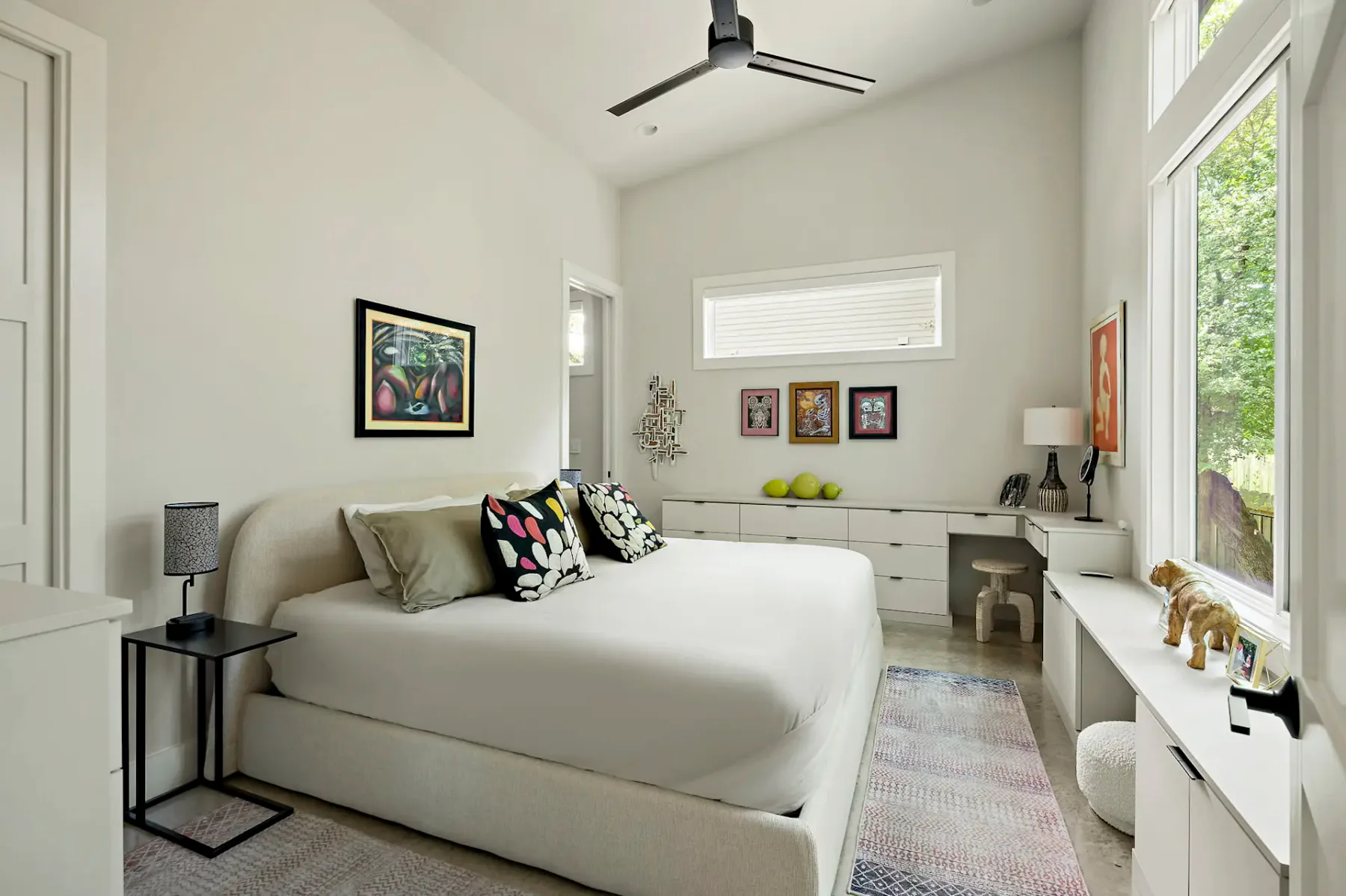 Dormitorio luminoso y moderno con una cama grande, almohadas coloridas, obras de arte, un ventilador de techo y una ventana grande con vista a la vegetación.