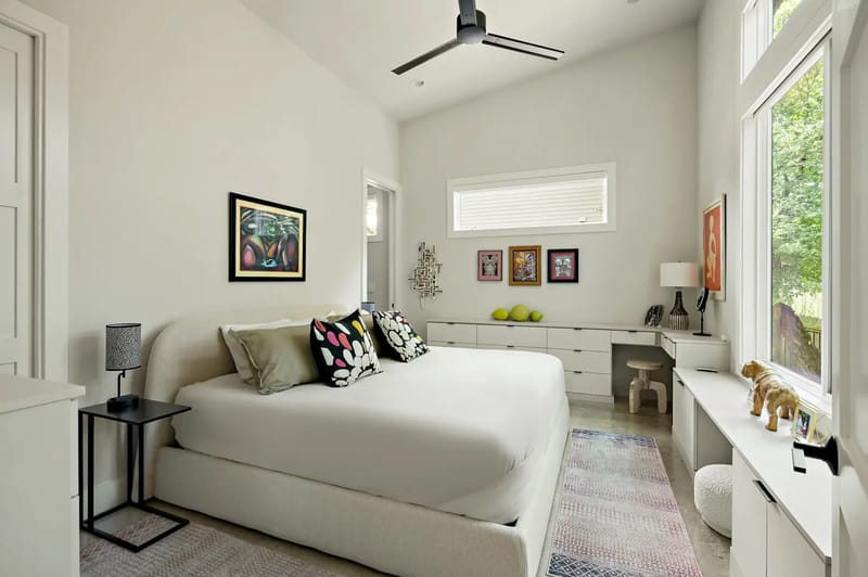 Bright, modern bedroom with a large bed, colorful pillows, artwork, a ceiling fan, and a large window overlooking greenery.