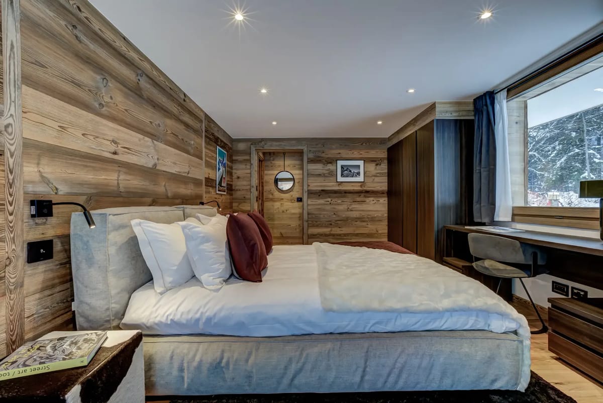 A cozy bedroom with wooden walls, a bed with white and burgundy pillows, a window with a view, and a small desk and chair.