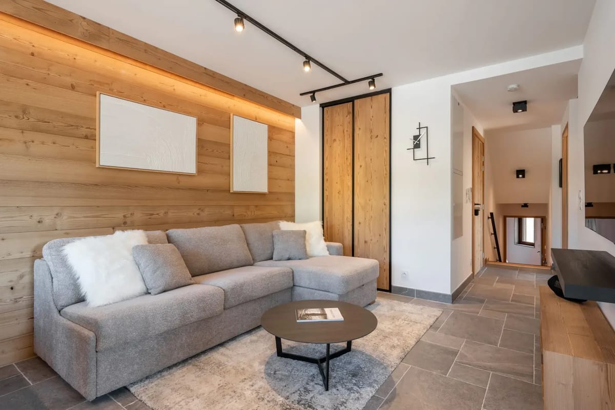Zona moderna de salón y comedor con paredes de madera, sofá modular gris, mesa de comedor de cristal, sillas negras, asientos tipo banco y una gran ventana que deja entrar luz natural.