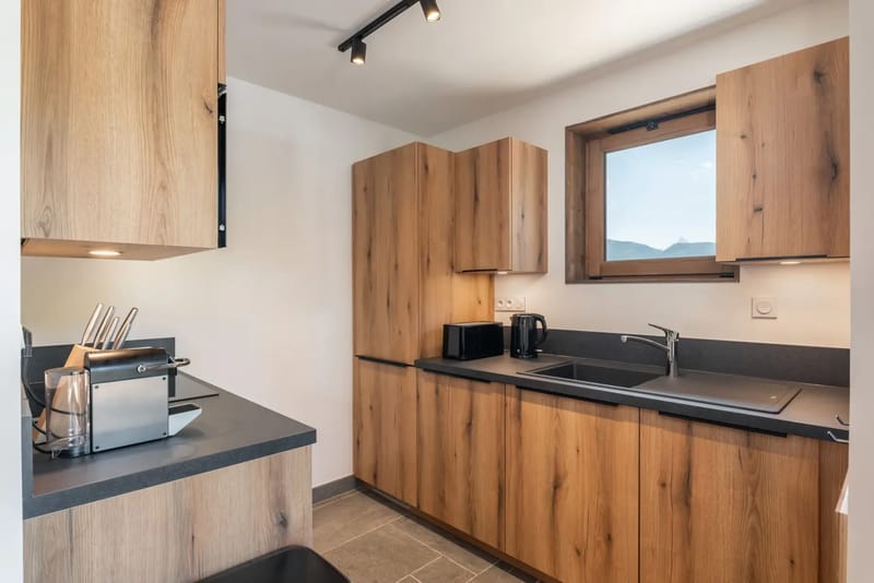 Modern kitchen with wood cabinets, black countertops, a sink, window, coffee maker, kettle, and knife block under ceiling and under-cabinet lights.