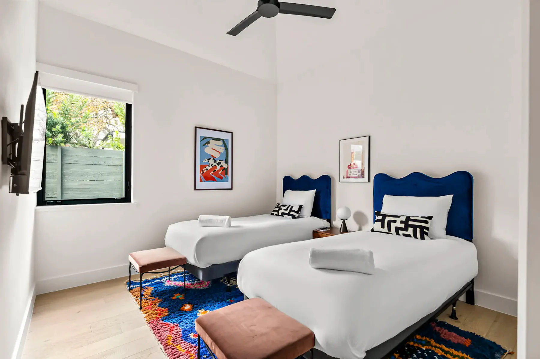 Dormitorio con dos camas individuales, una alfombra colorida, obras de arte enmarcadas, una ventana con vistas a la vegetación y un ventilador de techo.