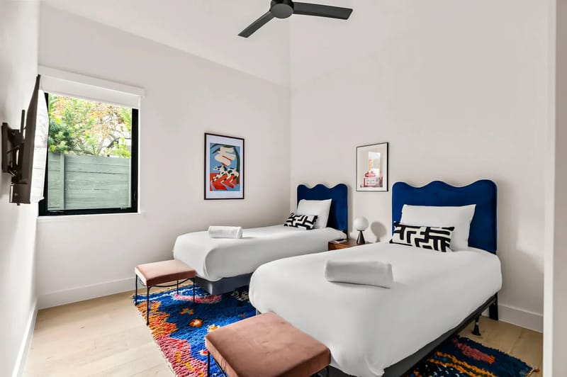 Bedroom with two twin beds, colorful rug, framed art, a window overlooking greenery, and a ceiling fan.