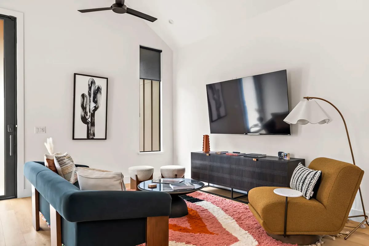 Modern living room with a blue sofa, mustard chair, mounted TV, cactus artwork, floor lamp, and geometric rug.