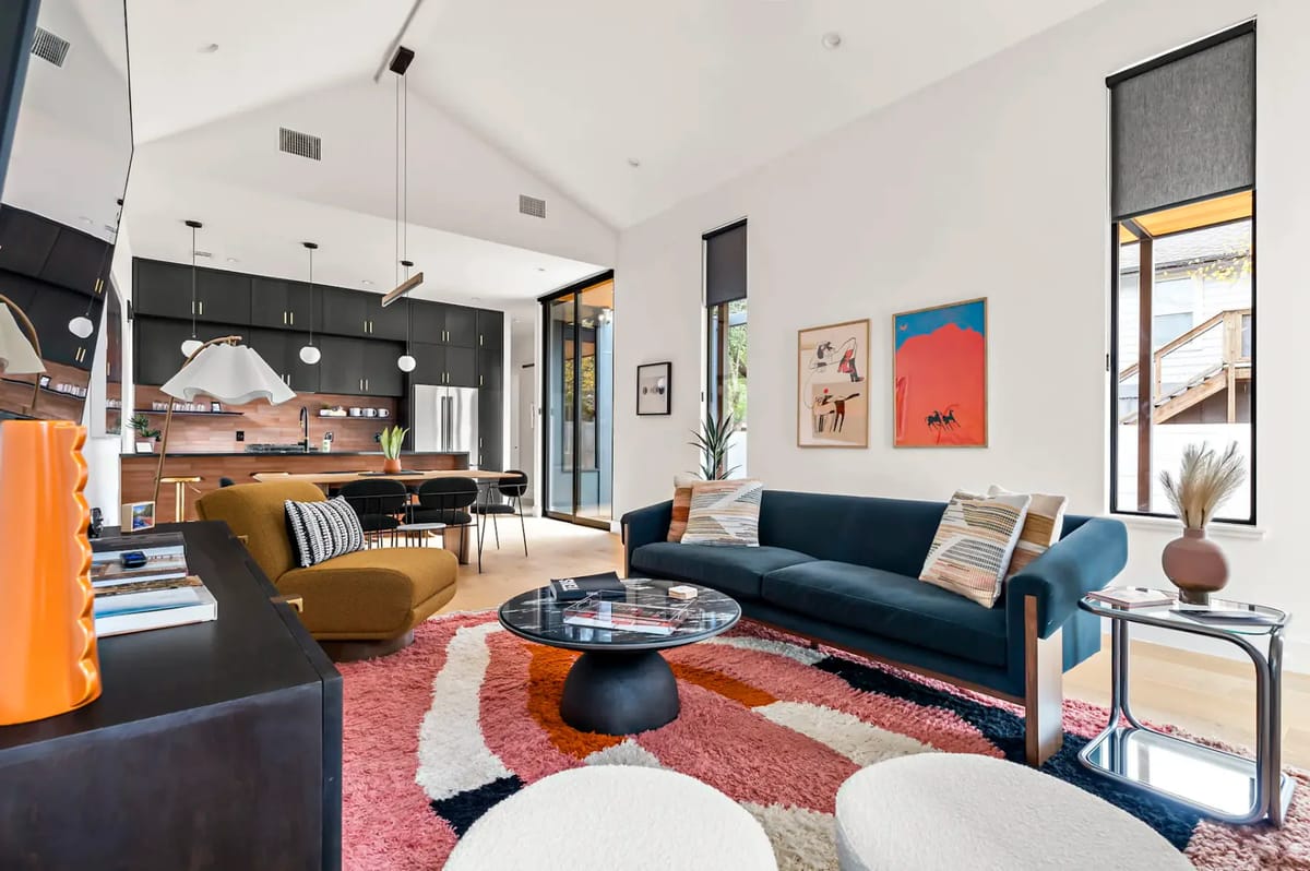 Modern living room with teal sofa, colorful rug, wall art, and a flat-screen TV; the open kitchen and dining area are visible in the background.