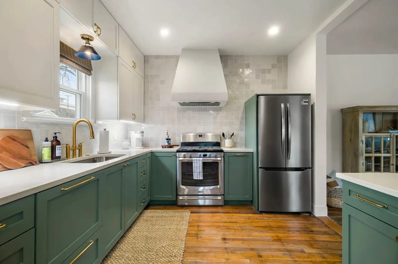 Cocina moderna con gabinetes verdes, electrodomésticos de acero inoxidable, encimeras blancas y piso de madera.