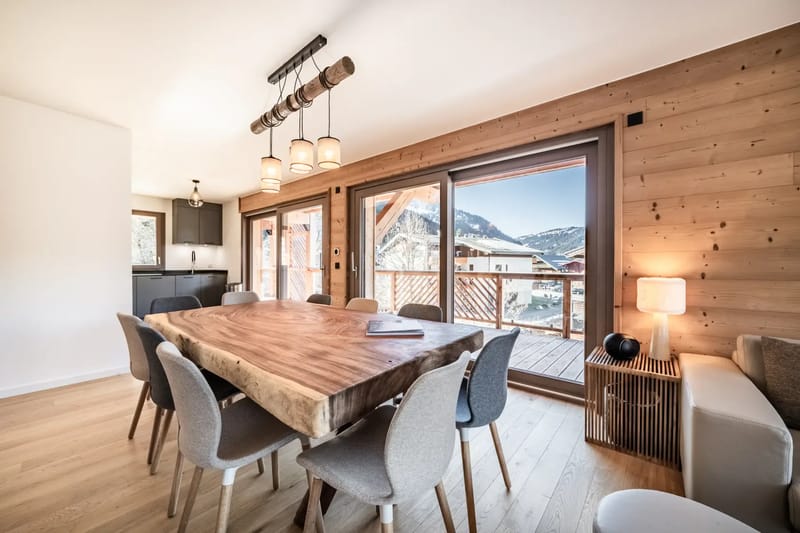 Un comedor espacioso con una mesa de madera, sillas variadas, paredes revestidas de madera, ventanas grandes con vistas a las montañas y una lámpara de araña moderna.