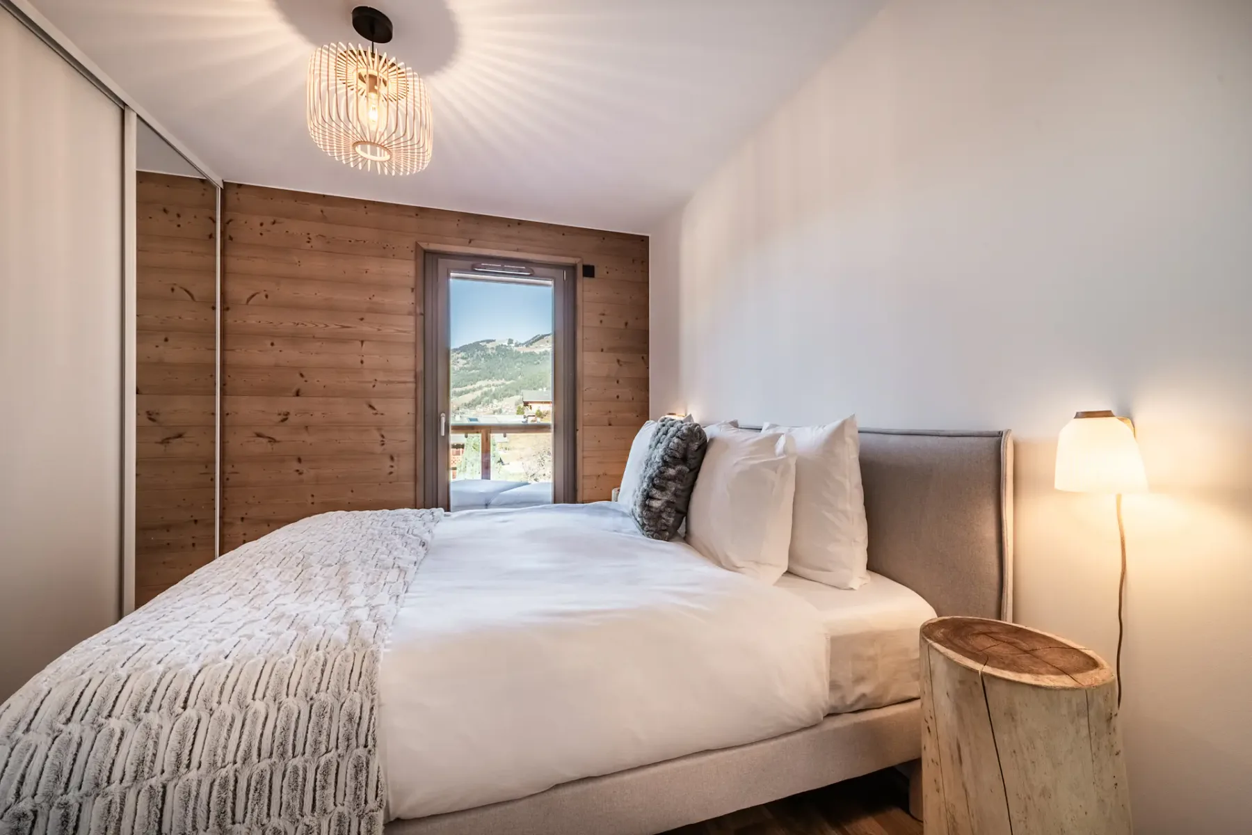 Dormitorio acogedor con una cama grande, una pared decorativa de madera, una lámpara de mesilla y vistas a las montañas desde la ventana. Una luminaria moderna en la parte superior.