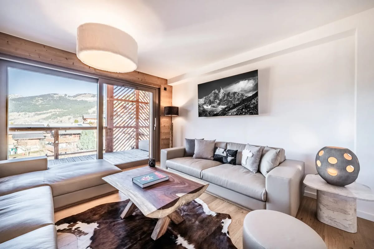 Modern living room with beige sofas, wooden walls, large window with mountain view, abstract art, and cozy lighting.