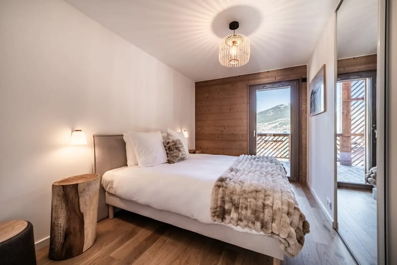 Dormitorio acogedor con una cama doble, paredes de madera, iluminación suave y una ventana grande con vistas a la montaña. La decoración rústica incluye una mesa auxiliar de madera.