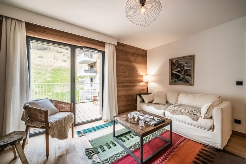 Acogedora sala de estar con un sofá blanco, alfombras de colores y un televisor montado en la pared. Una puerta corredera da a una zona exterior.