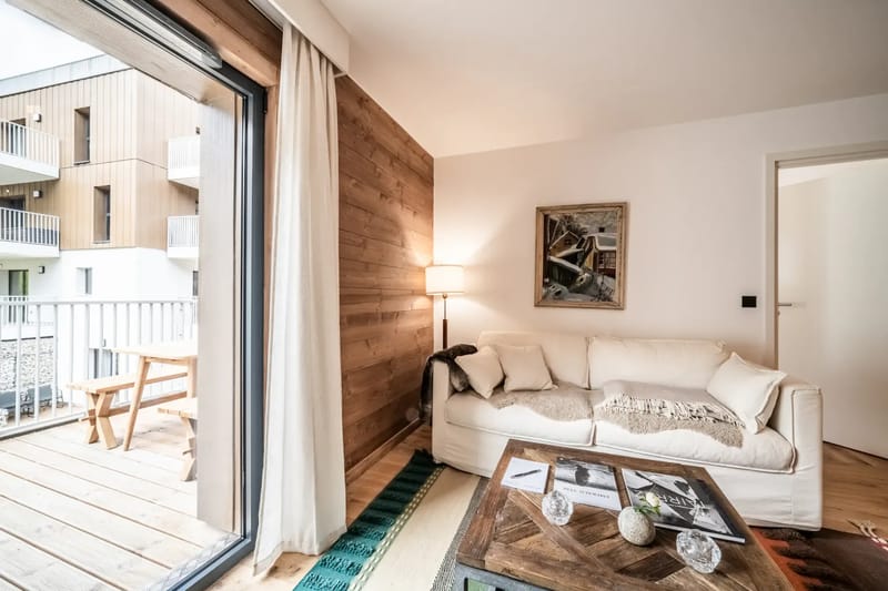 Acogedora sala de estar con un sofá blanco, alfombras de colores y un televisor montado en la pared. Una puerta corredera da a una zona exterior.
