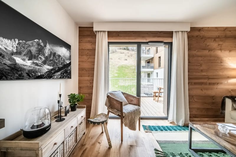 Acogedora sala de estar con un sofá blanco, alfombras de colores y un televisor montado en la pared. Una puerta corredera da a una zona exterior.