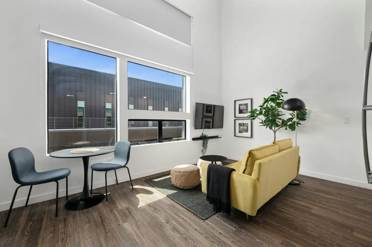 Modern living room with a yellow sofa, round table with chairs, large windows, floor lamp, and indoor plant on wooden flooring.
