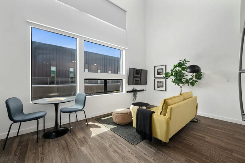 Sala de estar moderna con un sofá amarillo, mesa redonda con sillas, ventanas grandes, lámpara de pie y una planta de interior sobre un piso de madera.