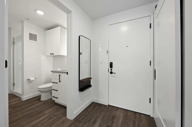 Baño moderno blanco con piso de madera, inodoro, mueble de lavabo con espejo y una entrada compacta.