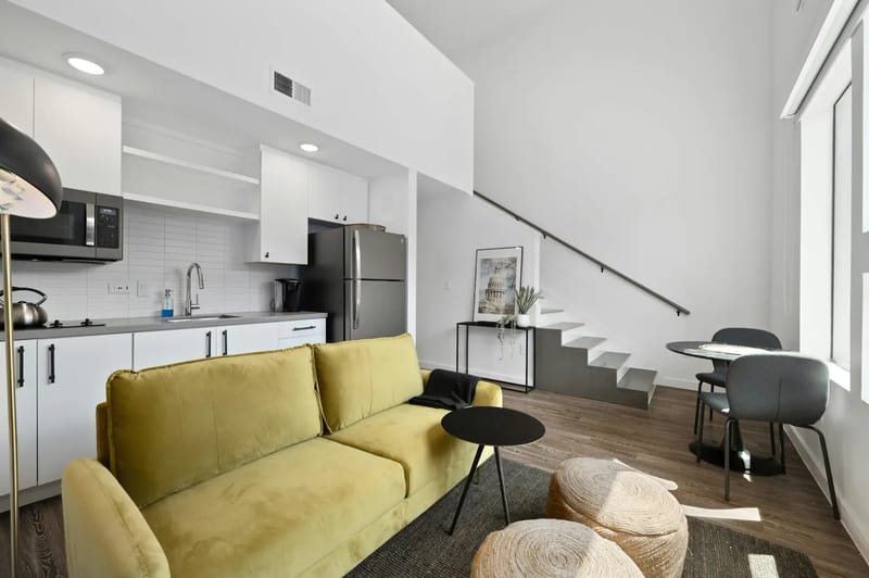 Apartamento tipo estudio moderno con techos altos, un sofá amarillo, cocina pequeña, un comedor pequeño y una escalera blanca junto a una pared luminosa.