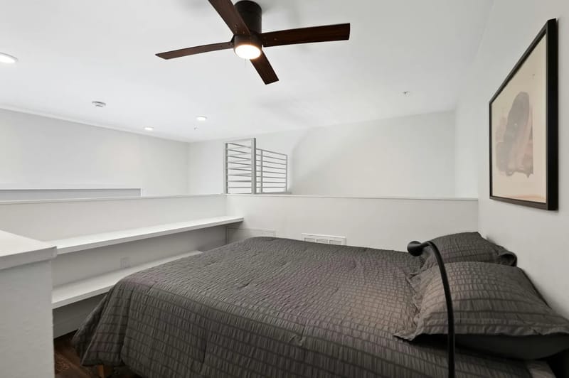 Dormitorio minimalista con una cama con colcha acolchada gris, paredes blancas, ventilador de techo e iluminación suave.