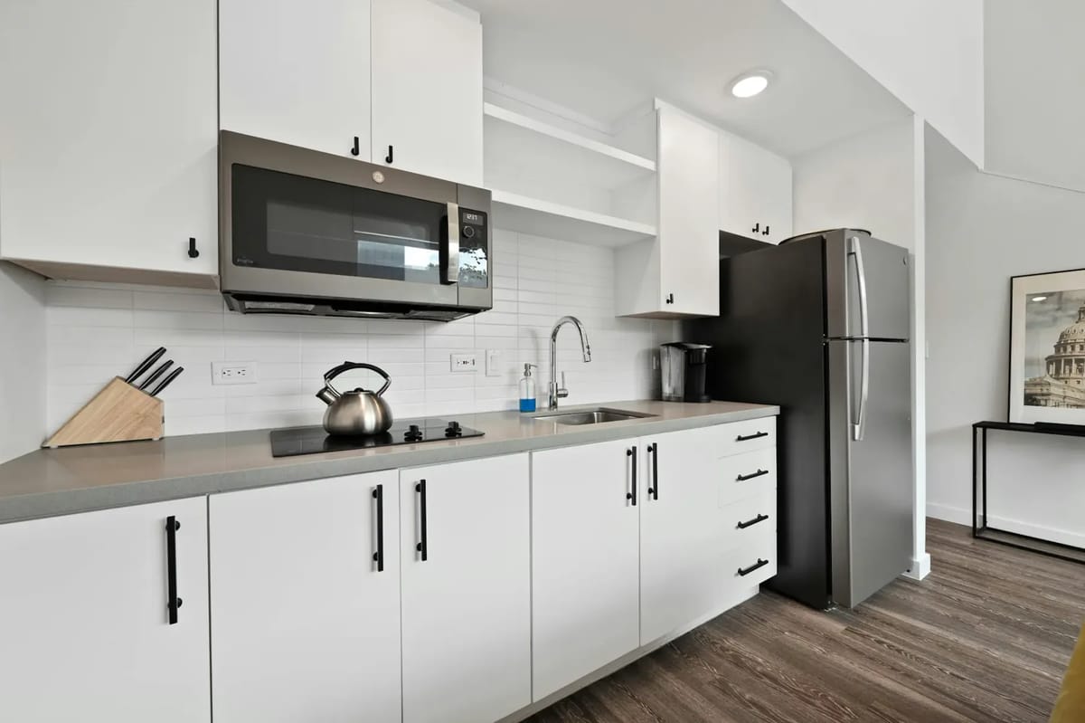 Modern kitchen with white cabinets, stainless steel appliances including a microwave and fridge, a kettle on the stovetop, and wooden flooring.