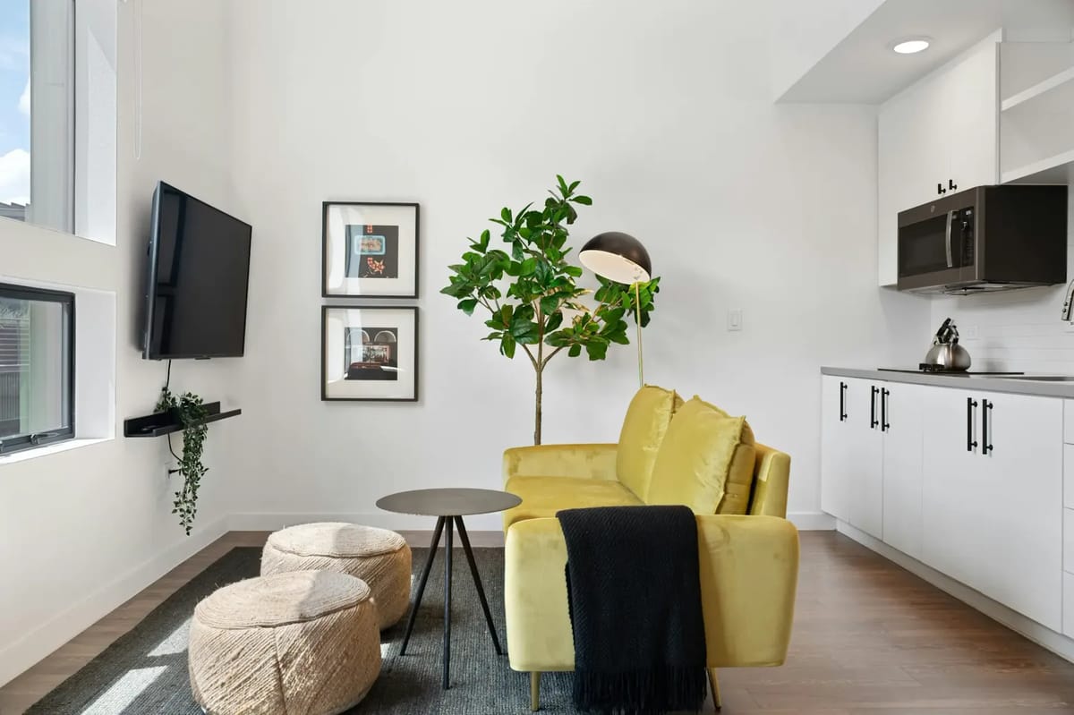 Modern living room with a yellow sofa, round table, ottomans, wall-mounted TV, plant, floor lamp, and a kitchen area with white cabinets.