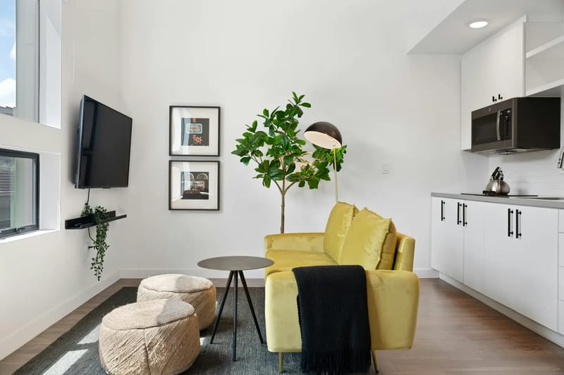 Sala de estar moderna con un sofá amarillo, mesa redonda, pufs, televisor montado en la pared, planta, lámpara de pie y un área de cocina con gabinetes blancos.