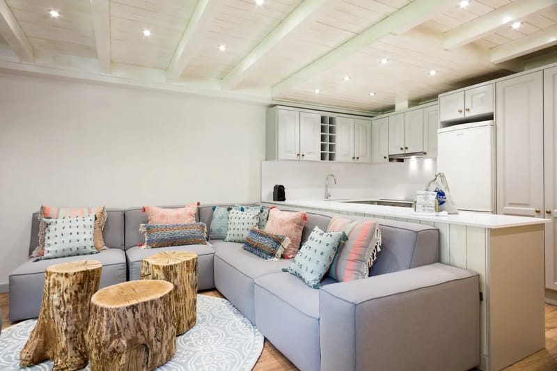A modern open-concept living and dining area with wooden accents. A gray sofa, wooden coffee table, and poufs are in the foreground, while a dining table with chairs and kitchen are in the background. Large windows provide natural light.