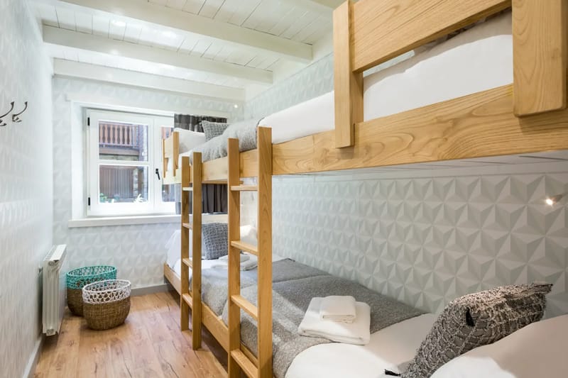 A cozy attic bedroom with a wooden ceiling and light blue walls. The room features a double bed with gray and white bedding, two lamps, a small wooden bench, and a skylight window.