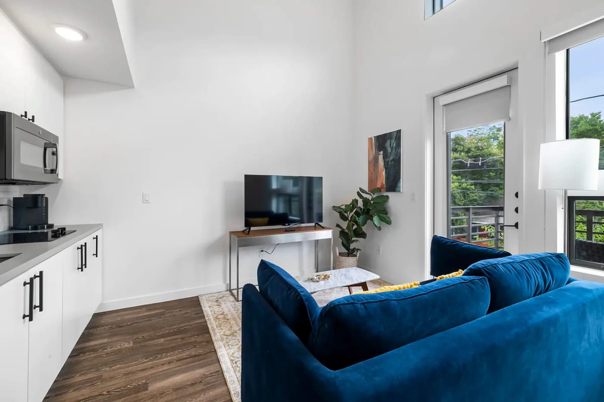 Sala de estar moderna con un sofá azul, televisor grande, una planta y una pequeña área de cocina. La puerta de vidrio da a un balcón con vistas a la vegetación exterior.