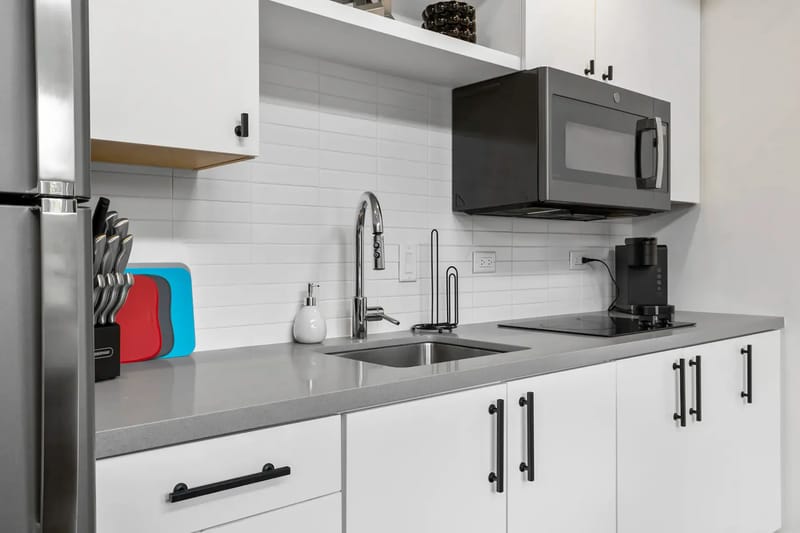 Cocina moderna con gabinetes blancos, encimera gris, fregadero, juego de cuchillos, microondas, cafetera y tablas de cortar frente a un salpicadero de azulejos blancos.