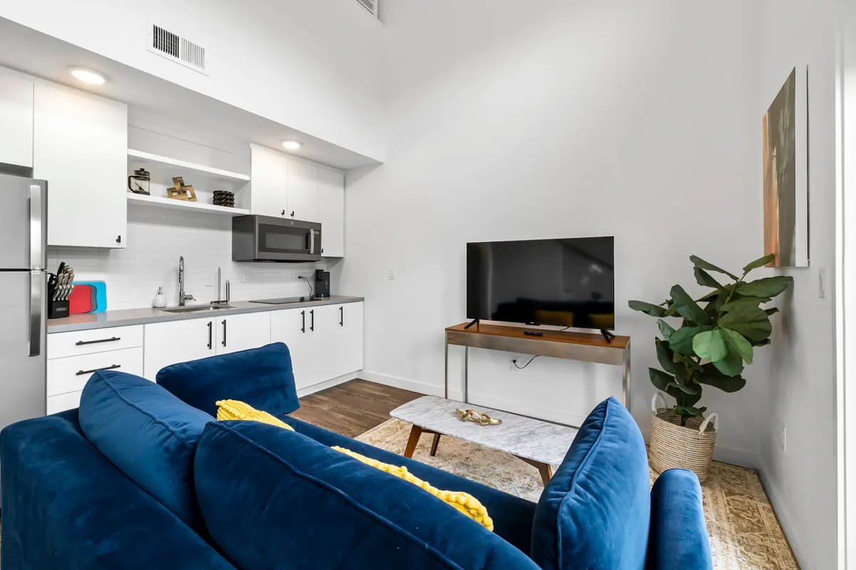 Sala de estar moderna con un sofá azul, cojines de colores, televisor sobre un mueble de madera, ventanas grandes y una planta. La puerta da a un balcón con vista a la vegetación.