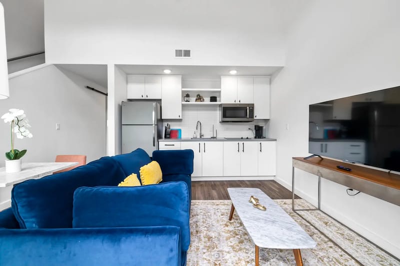 Sala de estar moderna con sofá azul, cojín amarillo, cocina pequeña blanca, televisor y mesa de centro de mármol sobre una alfombra estampada.
