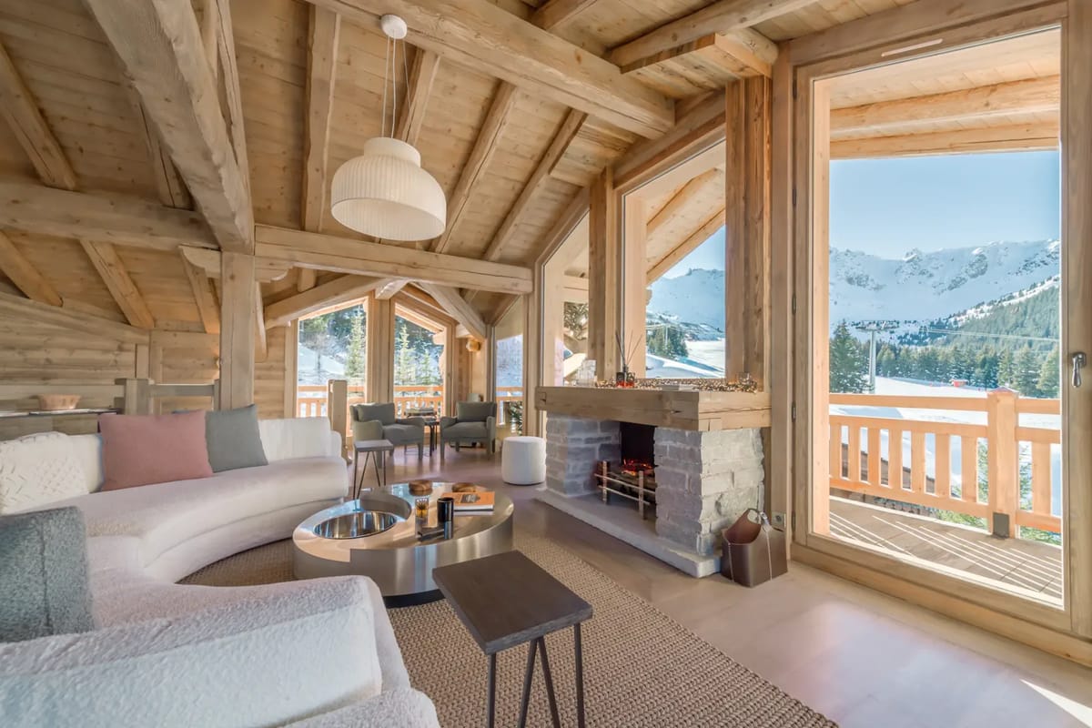 Acogedor interior de chalet de madera con un sofá curvo, chimenea y grandes ventanas con vistas a montañas nevadas y a un balcón de madera.