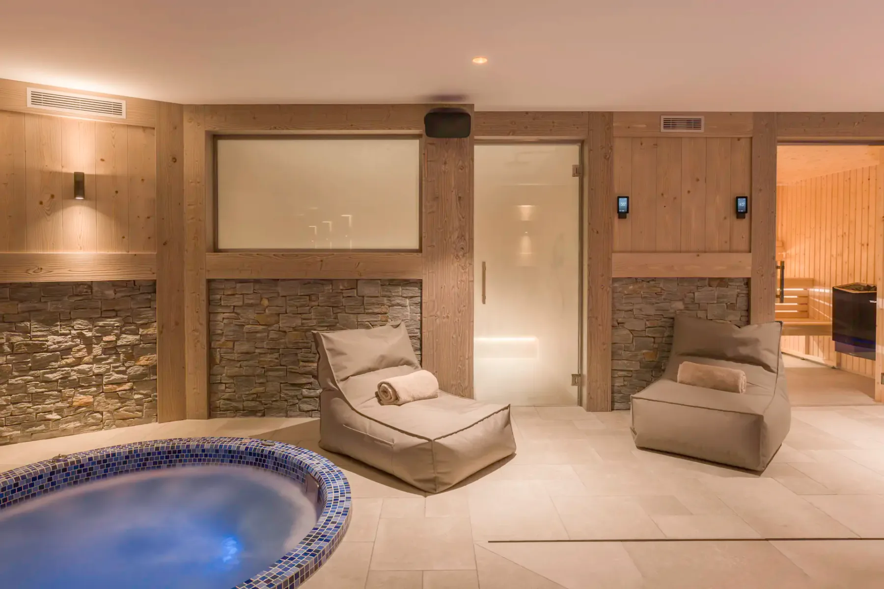 Sala de spa con una bañera de hidromasaje redonda, tumbonas, paredes de piedra y madera, y una sauna al fondo.