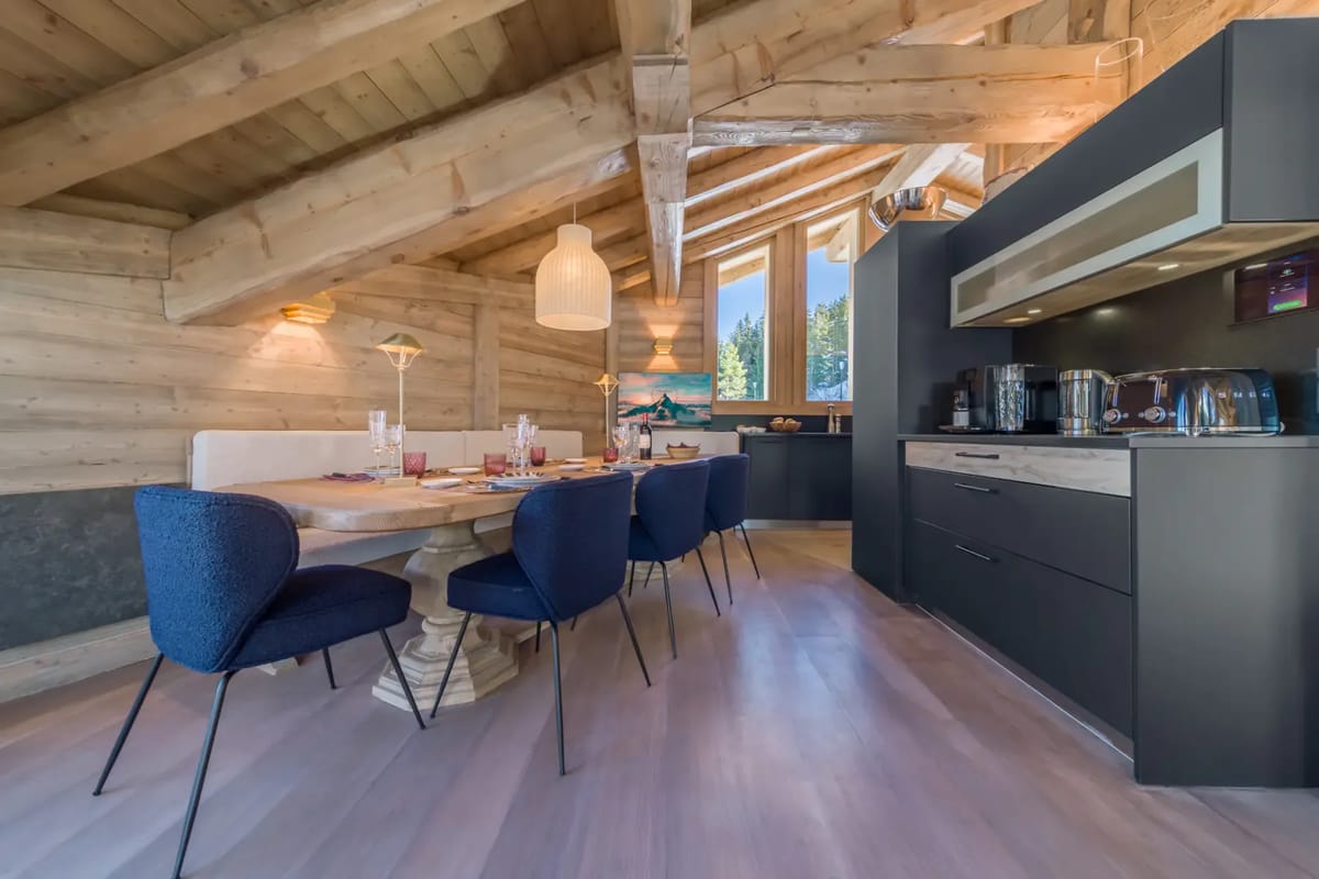 Acogedora zona de comedor con vigas de madera, una mesa puesta, sillas azules y electrodomésticos modernos bajo un techo inclinado con grandes ventanas.