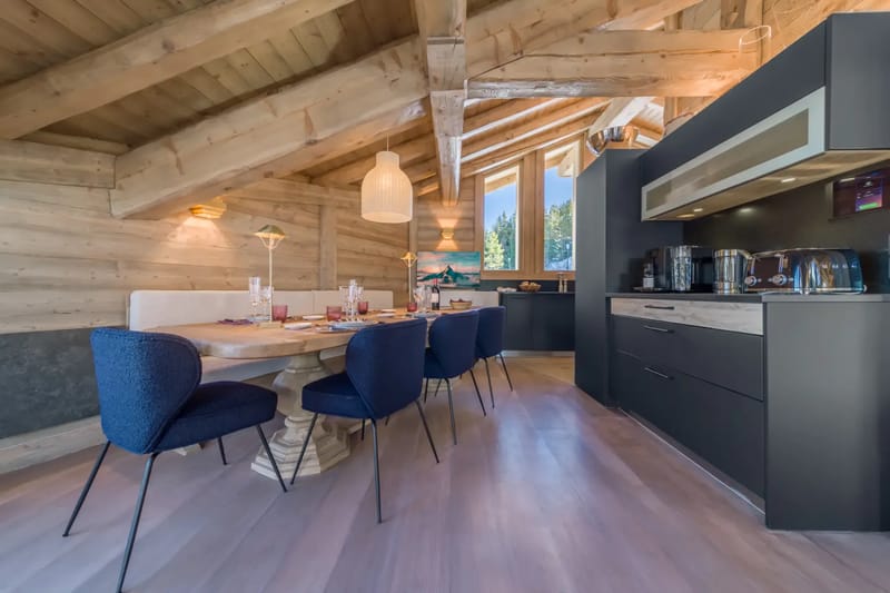Cozy dining area with wooden beams, a set table, blue chairs, and modern kitchen appliances under a sloped ceiling with large windows.