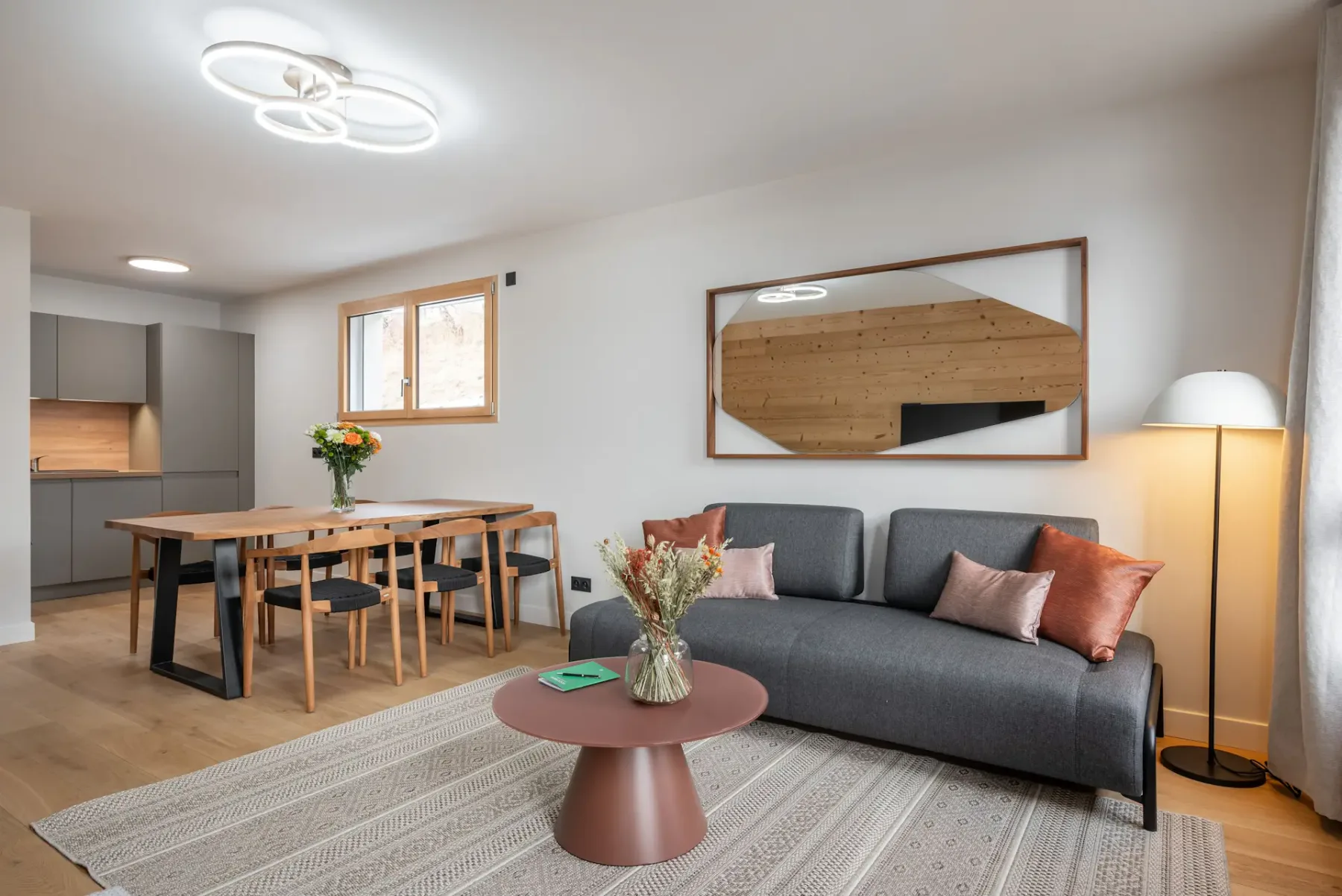 Salón moderno con paredes de madera, una gran mesa de comedor, sillas, un sofá gris y un televisor montado en la pared. Las grandes ventanas muestran un paisaje nevado.