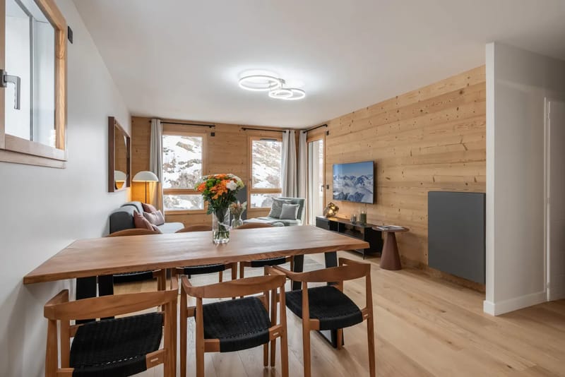 Salón moderno con paredes de madera, una gran mesa de comedor, sillas, un sofá gris y un televisor montado en la pared. Las grandes ventanas muestran un paisaje nevado.