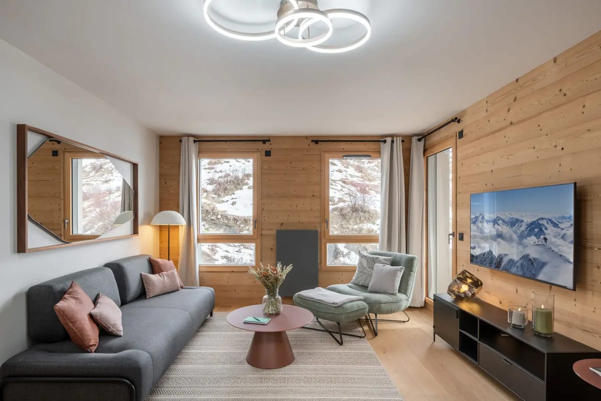 Modern living room with wood accents, a grey sofa, chairs around a wooden dining table, a cozy armchair, and large windows with a snowy view.