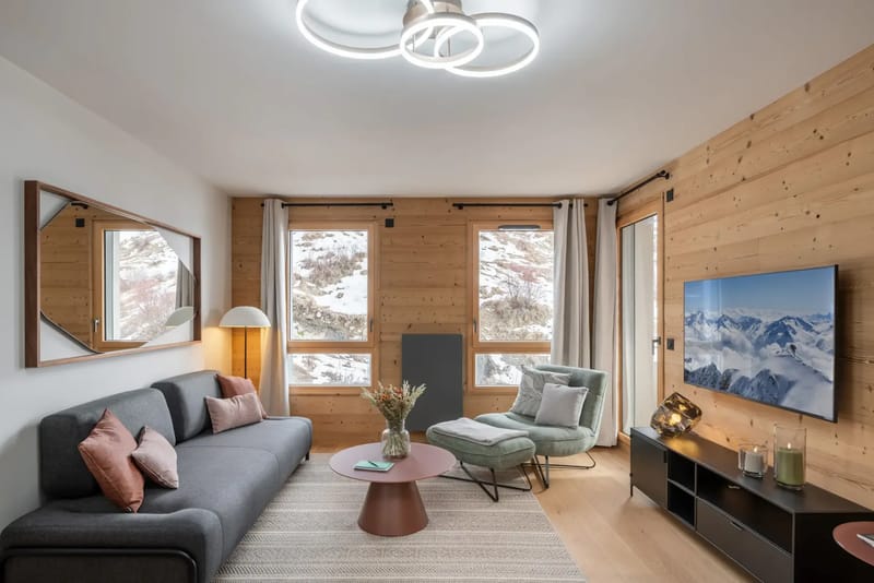 Salón moderno con detalles de madera, un sofá gris, sillas alrededor de una mesa de comedor de madera, un sillón acogedor y grandes ventanas con vistas a un paisaje nevado.