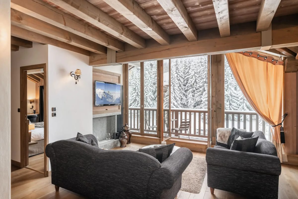 Salón con vigas de madera, grandes ventanas, sofás grises, una mesa de centro redonda y una zona de comedor.