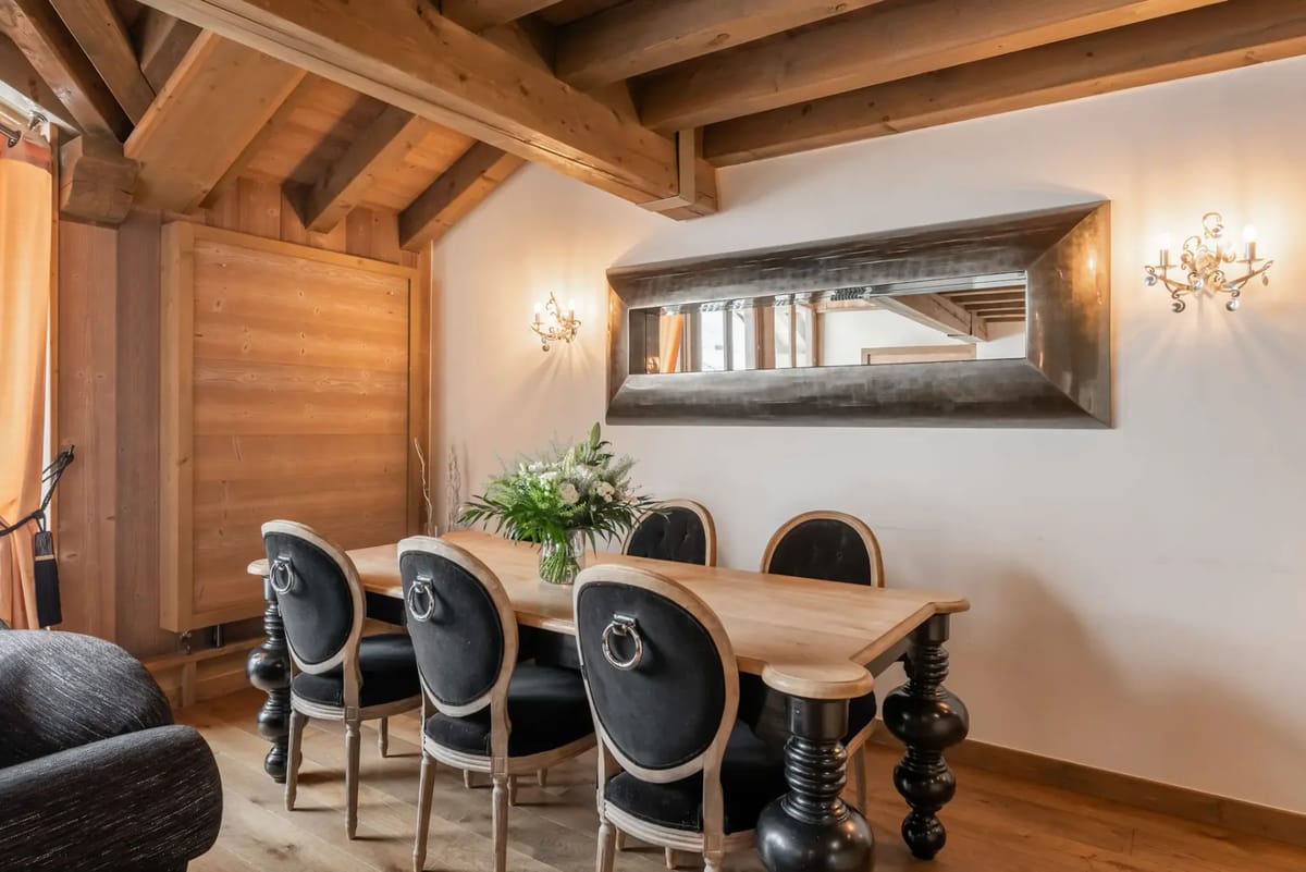 Comedor rústico con una mesa de madera, sillas negras acolchadas, un espejo grande y vigas de madera vistas.