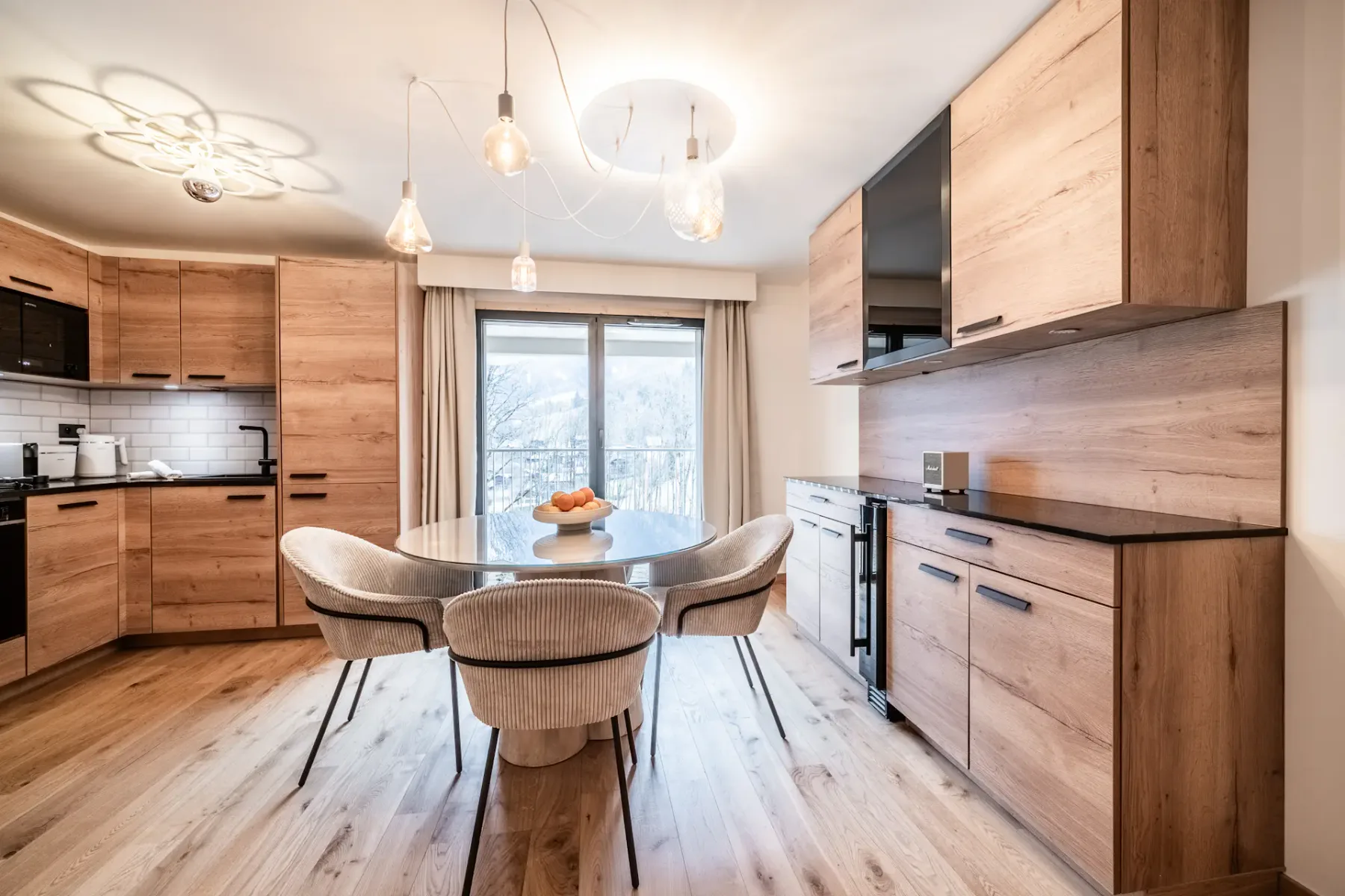 Modern kitchen with wood cabinets, round table with chairs, and a fruit bowl. Large window with beige curtains offers a mountain view. Pendant lights hang from the ceiling.
