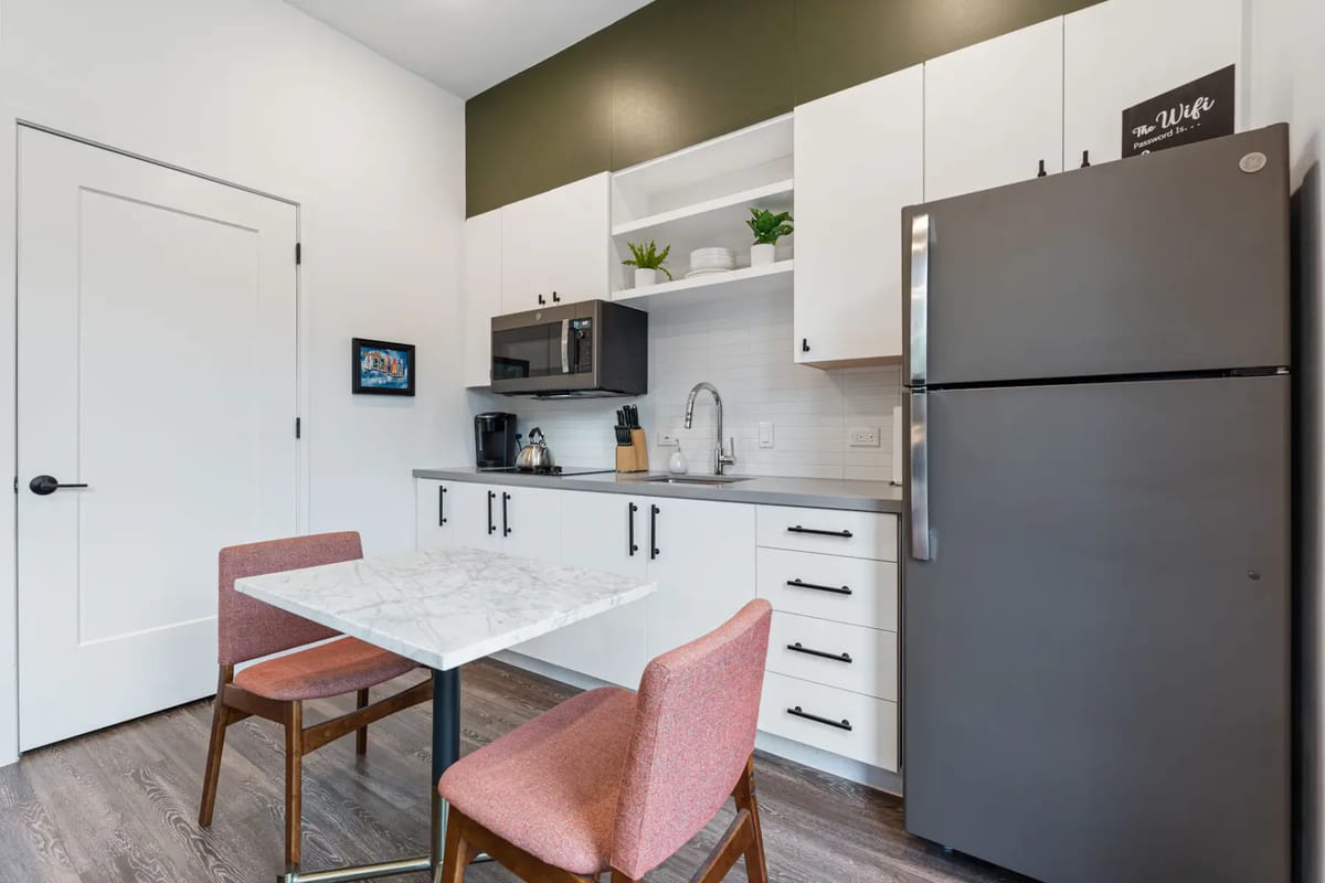 Cocina compacta con una mesa de mármol, dos sillas rosas, gabinetes blancos, un refrigerador gris, microondas y una pared de acento verde.