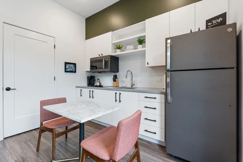 Cocina compacta con una mesa de mármol, dos sillas rosas, gabinetes blancos, un refrigerador gris, microondas y una pared de acento verde.