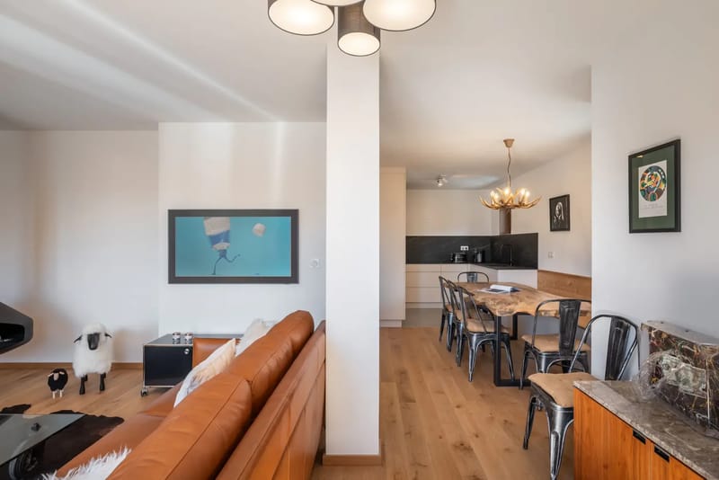 Zona de comedor con una mesa de madera, cuatro sillas metálicas, un libro abierto, una lámpara de araña con forma de astas, obras de arte en la pared y vistas a una sala de estar con grandes ventanas.