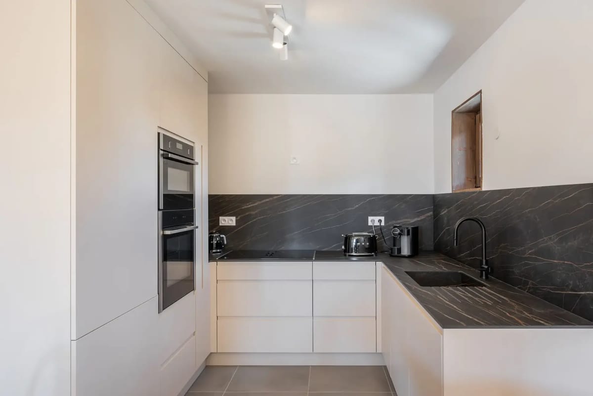 Cocina moderna con muebles claros, horno integrado, encimeras y salpicadero de mármol negro, fregadero y varios pequeños electrodomésticos sobre la encimera.