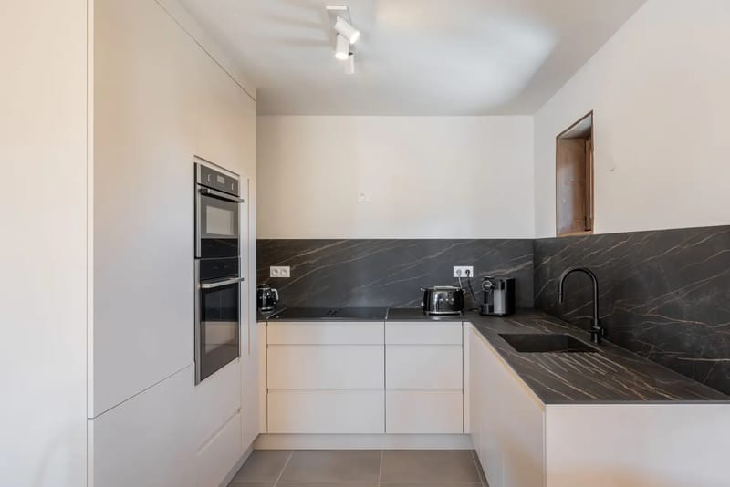 Cocina moderna con muebles claros, horno integrado, encimeras y salpicadero de mármol negro, fregadero y varios pequeños electrodomésticos sobre la encimera.