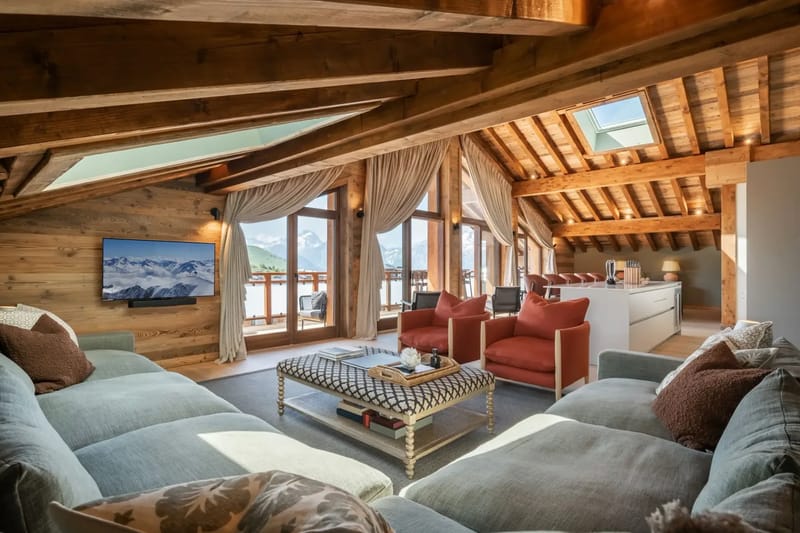 Acogedor salón abuhardillado con vigas de madera, grandes ventanas, un televisor y vistas a las montañas.