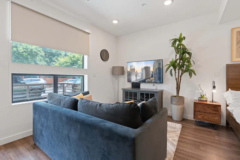 Modern living room with a blue sofa, TV, large window, and adjacent bed area.