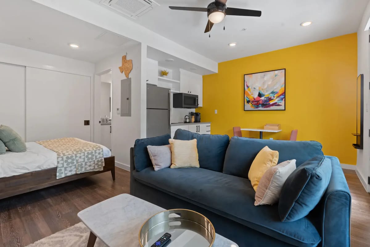 Modern studio apartment with a blue sofa, yellow accent wall, bed, small dining area, and kitchen.